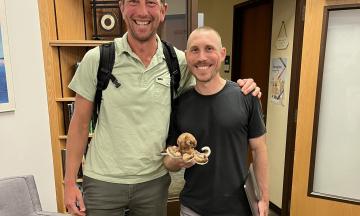 Photo of Fletcher Beaudoin and Paul Loikith standing next to each other. Paul is holding a stuffed octopus. 