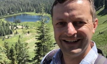 Photo of Max Nielsen-Pincus, the background is a forest landscape with a blue lake 