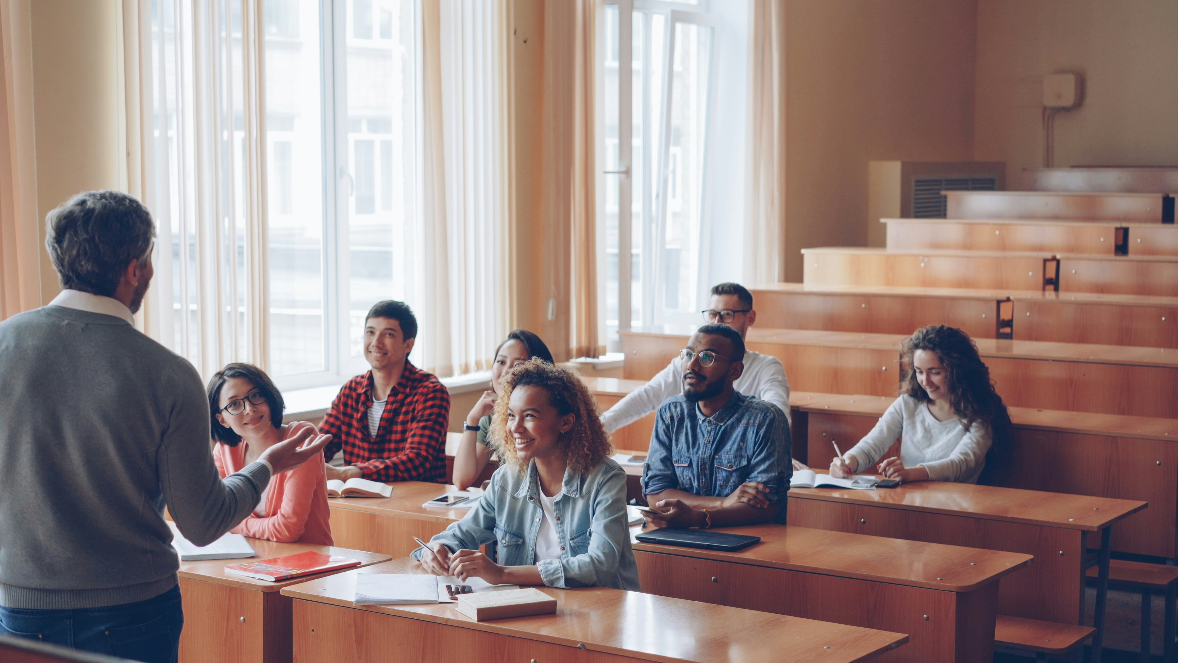 Students sit in a classroom at wooden desks, facing the front of the room.