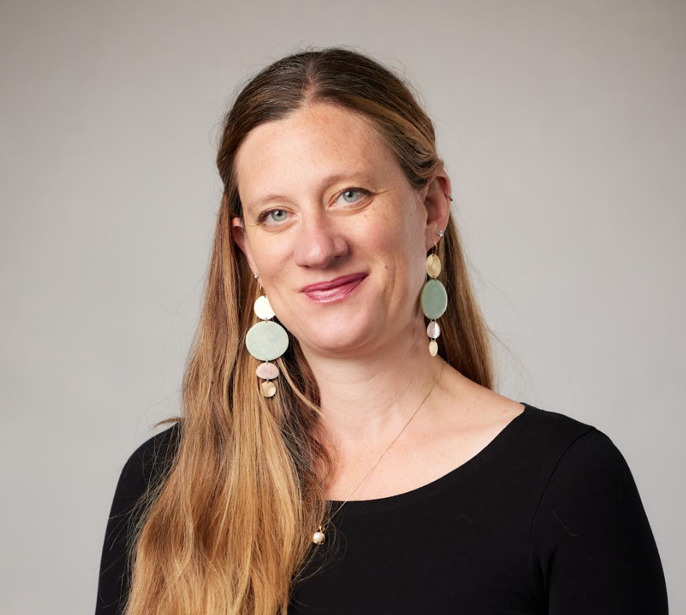 Kathleen smiles with mouth closed, has brown hair past their shoulders, wearing long earrings with stacked circles, and black top.