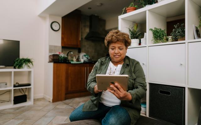 Woman with brown hair and green jacket holds an iPad while sitting on her couch