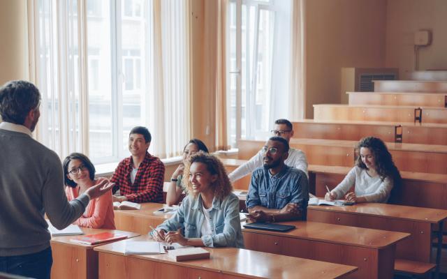 Students sit in a classroom at wooden desks, facing the front of the room.