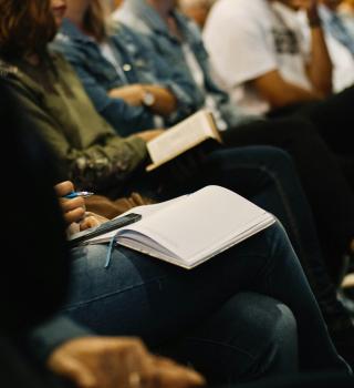 People sit close together in a row with several notebooks open in their laps.