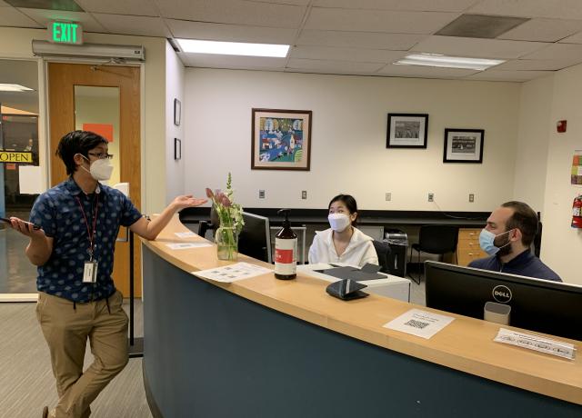 Two staff members sit at the front desk and welcome an international student standing at the front desk.