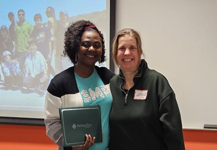 Student smiles as they hold certificate next to the teacher in front of a classroom.