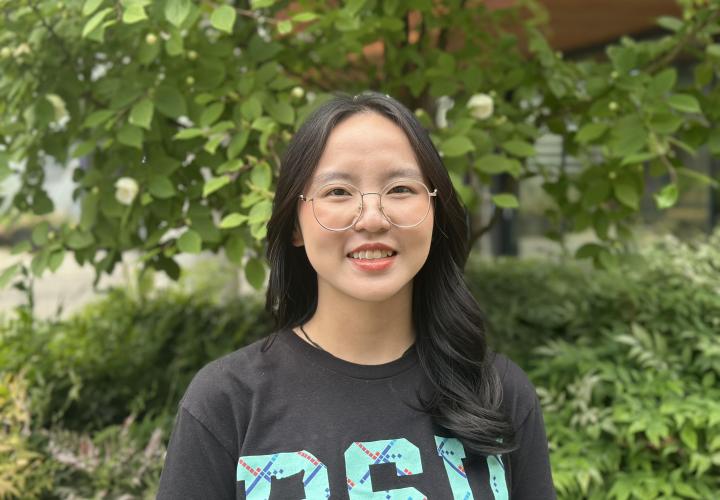 Female student poses for a headshot outside with greenery in the background.