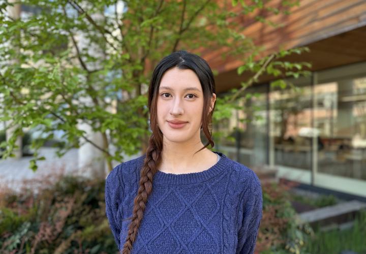 Female student poses for a headshot outside with greenery in the background.