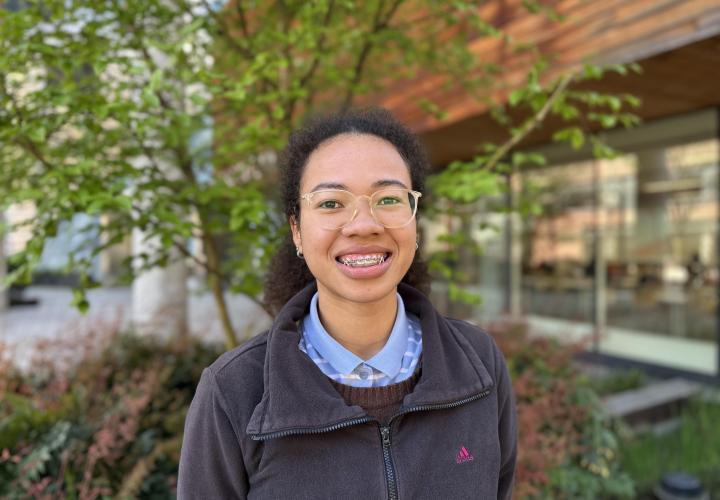 Female student poses for a headshot outside with greenery in the background.