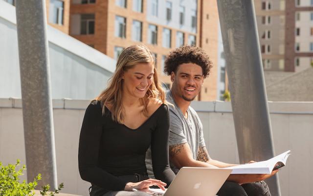 A female and male student sit next to each other outside. The female student is looking at her laptop. The male student is holding a text book.