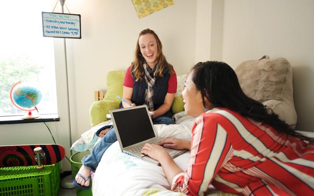 Two female students in their dorm room having a conversation while sitting on a bed.