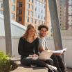 A female and male student sit next to each other outside. The female student is looking at her laptop. The male student is holding a text book.