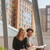 A female and male student sit next to each other outside. The female student is looking at her laptop. The male student is holding a text book.
