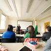 A group of students sit in a classroom with an instructor at the front. One student raises her hand.