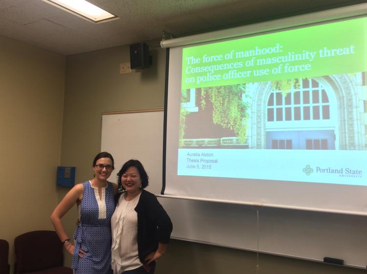 Aurelia Alston and J. Katherine Lee standing in front of the projected presentation slides for Aurelia's thesis proposal meeting. The slide reads: "The force of manhood: Consequences of masculinity threat on police officers use of force. Aurelia Alston Thesis Proposal. June 5th, 2015