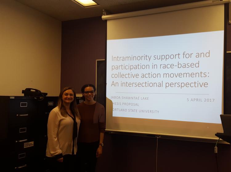 Dr. Kahn and Jaboa Lake standing in front of the projected presentation slides for Jaboa's thesis proposal meeting. The slide reads: "Intraminority support for and participation in race-based collective action movements: An intersectional perspective. Jaboa Shawntaé Lake Thesis Proposal. Portland State University, April 5th, 2017." 