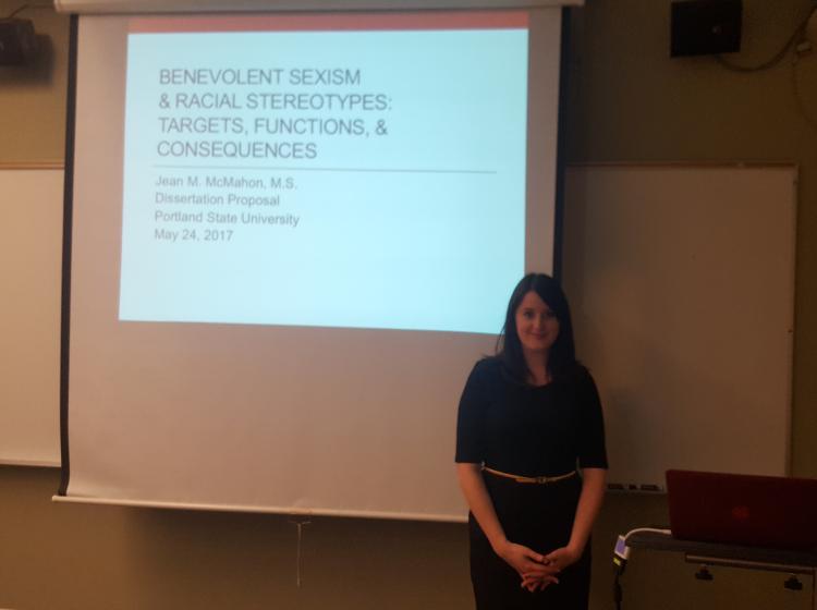Jean McMahon standing in front of the projected presentation slides for her dissertation proposal meeting. The slide reads: "Benevolent Sexism and Racial Stereotypes: Targets, Functions, and Consequences. Jean M. McMahon, M.S., Dissertation Proposal. Portland State University, May, 24th, 2017."