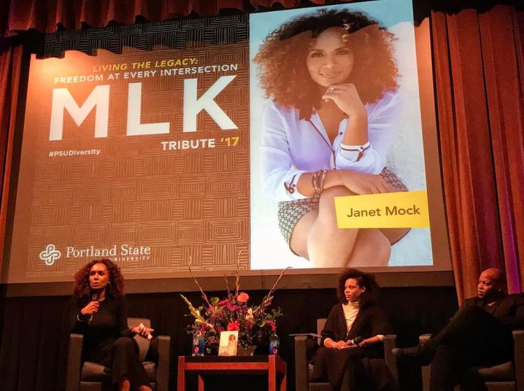 Photo of GRASP Lab member Jaboa Lake on a panel with Janet Mock and one unidentified speaker at the MLK Tribute event at Portland State University in 2017. The three are sitting on a stage in chairs. The slide behind them reads: "Living the legacy: Freedom at every intersection. MLK Tribute '17. #PSUdiversity. Janet Mock." The slide also features a photo of Janet Mock with her legs crossed and chin resting on one hand.