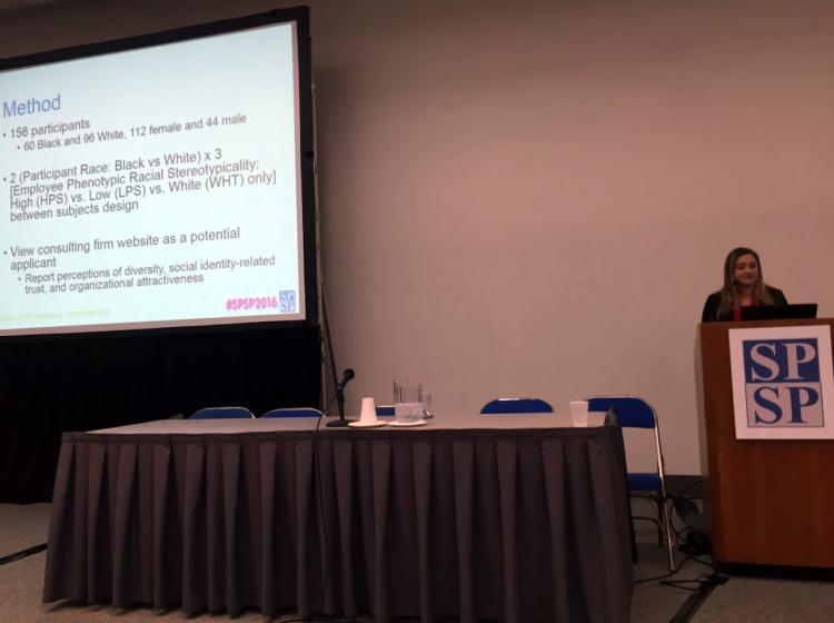 Photo of Dr. Kahn standing at a podium in front of an audience giving a talk with a large projector screen behind her. The screen behind her shows the methods for the research she is presenting.