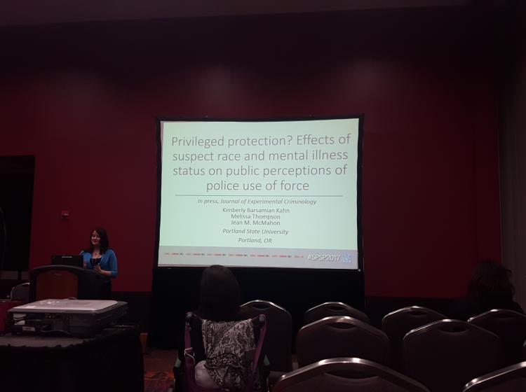 Photo of GRASP Lab member Jean McMahon standing at a podium in front of an audience giving a talk with a large projector screen behind her. The talk is titled: "Privileged Protection? Effect of suspect race and mental illness status on public perceptions of police use of force"