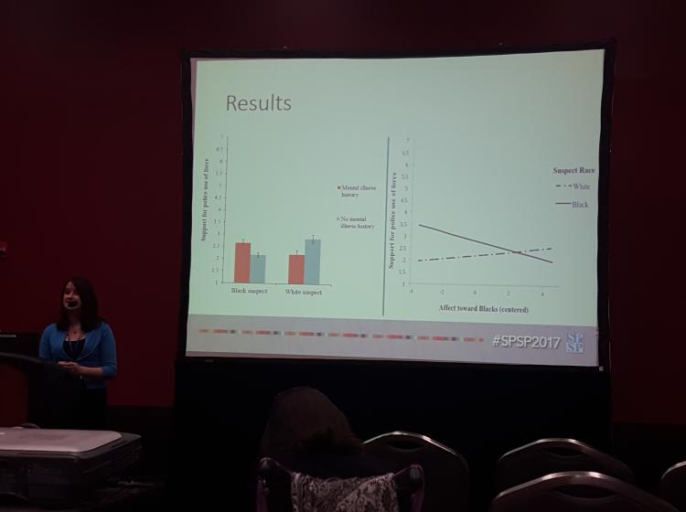 Photo of GRASP Lab member Jean McMahon standing at a podium in front of an audience giving a talk with a large projector screen behind her. The slide behind her shows a barchart and well as a line graph with two lines crossing indicating an interaction between two variables.