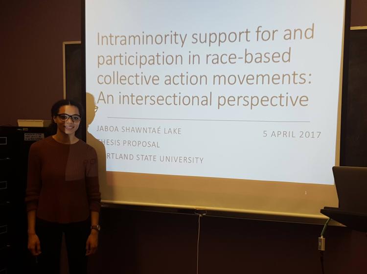Jaboa Lake standing in front of her title slide for her thesis proposal in April of 2017. The title on the slide reads "Intraminority support for and participation in race-based collective action movements: An intersectional perspective"