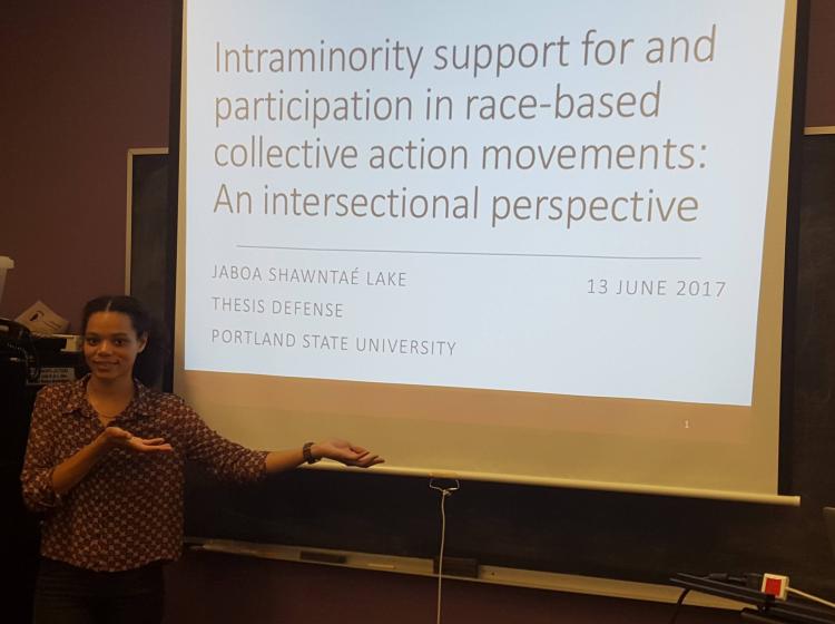 Jaboa Lake standing in front of her title slide for her thesis defense in June of 2017. The title on the slide reads "Intraminority support for and participation in race-based collective action movements: An intersectional perspective"