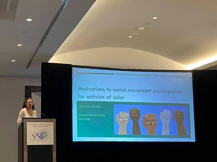 Jaboa Presentation at the 2022 Annual SPSSI Conference. Jaboa is standing behind a podium speaking with her title slide behind her. The title reads "Motivations to social movement participation for activists of color"