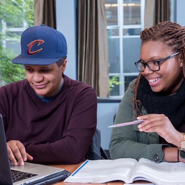 Two students sitting at a table with a laptop and book in front of them.