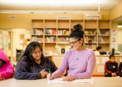 Two women at a table, one writing in a workbook