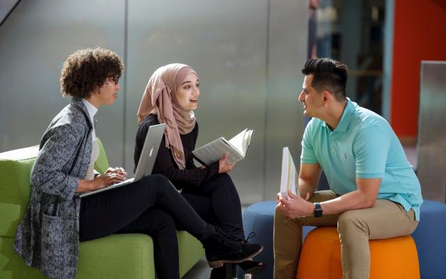 Three students conversing on colorful seats, one with laptop; two with books