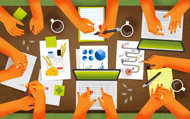Top-down view of a group of people collaborating at a table with paperwork, coffee mugs, laptops, pens and other stationary