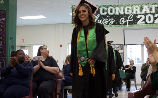 A graduate in a cap and gown and green sash walks forward smiling as an audience claps under a graduation banner
