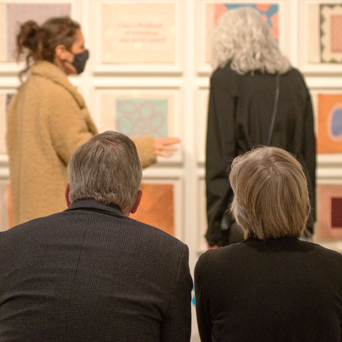 two seated people look on as a gallery attendant talks to a vistor about artwork on the wall. 