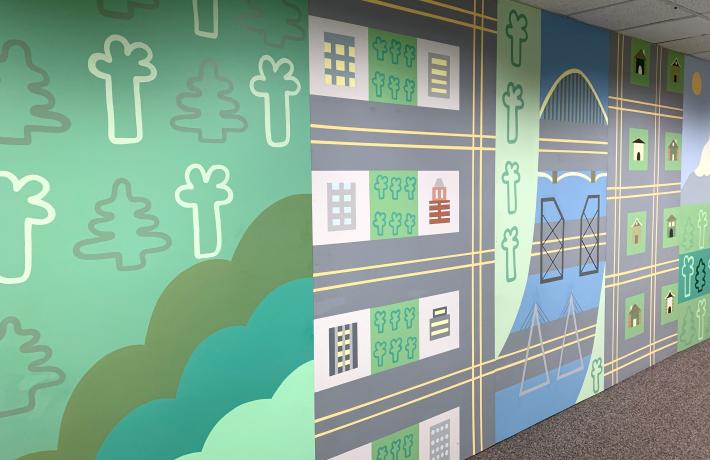 A long floor-to-ceiling mural in a carpeted hallway. The mural is a stylized depiction of Portland with trees, buildings, streets and river.
