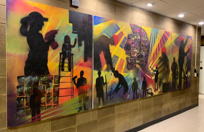 Full mural is shown on a brick wall in a hallway. It consists of four panels featuring abstract compositions showing workers with tools.