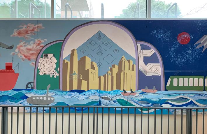 A mural above a stairwell, an Art Deco-style composition featuring a stylized Portland skyline at center, also featuring a river, ship, airplane and rocket.