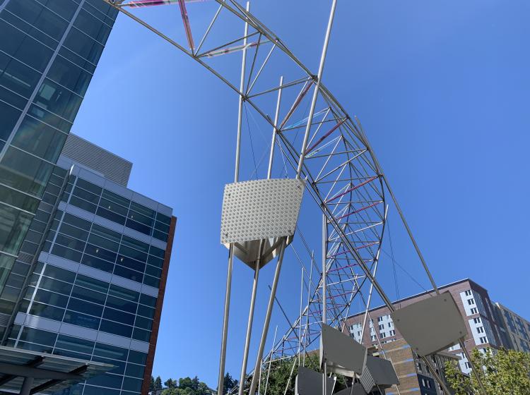 Close-up of a large metal sculpture stretching over the plaza in front of the Engineering Building, in an arc shape consisting of narrow rods and reflective bars.