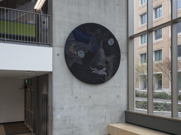 A large round multimedia artwork hangs on a wall in a well lit atrium area. The artwork is black and decorated in sequins and fabric, vaguely resembling stars and galaxies.