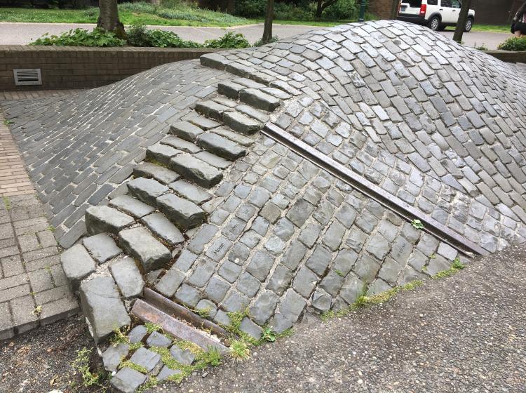 A mound of cobblestones, with a path of raised bricks leading over it. Words are etched in some stones, and pieces of railroad track embedded in the mound.
