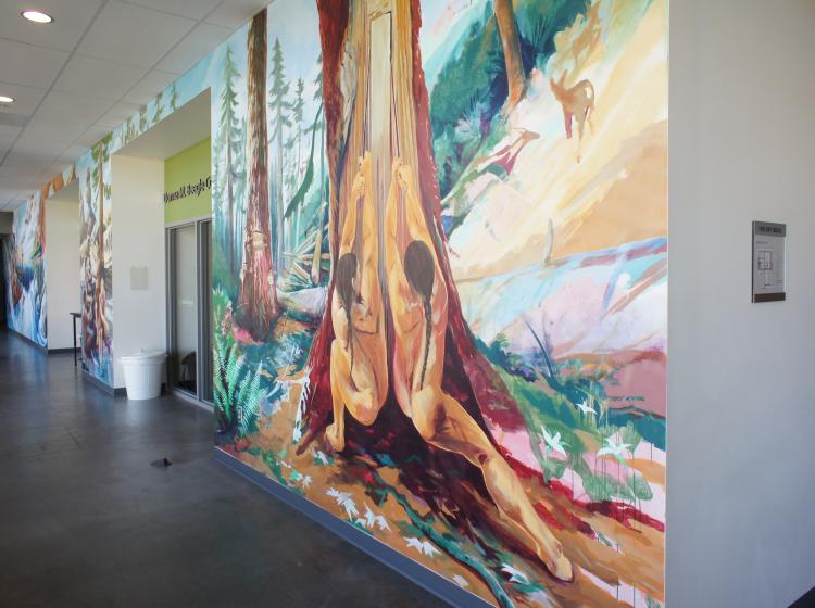 Angled shot of a section of the mural in the hallway. This section of the mural shows Native people stripping wood from a tree.