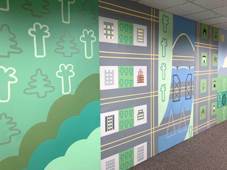 A long floor-to-ceiling mural in a carpeted hallway. The mural is a stylized depiction of Portland with trees, buildings, streets and river.