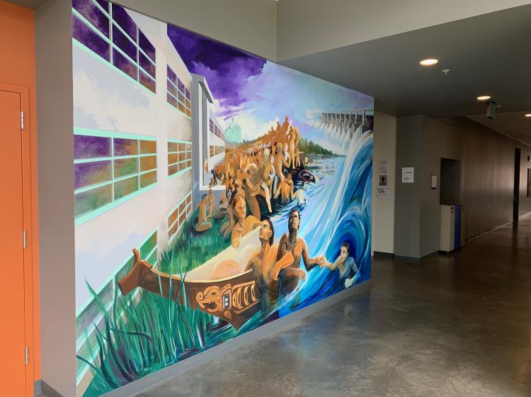Another section of the mural, in an adjacent hallway, that depicts Native people outside a building, surrounded by water rushing through a dam.