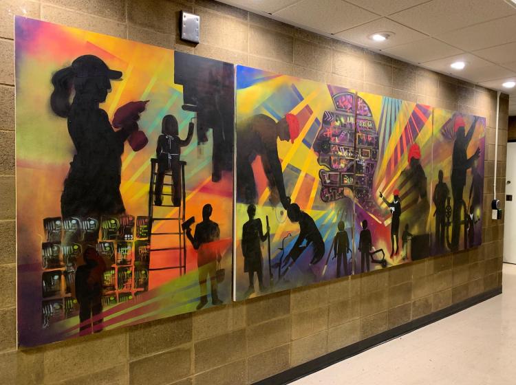 Full mural is shown on a brick wall in a hallway. It consists of four panels featuring abstract compositions showing workers with tools.
