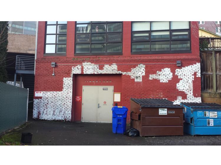 The brick wall is shown before the mural was painted. It is colored mostly red but with patches of white. There are dumpsters in front of the wall.