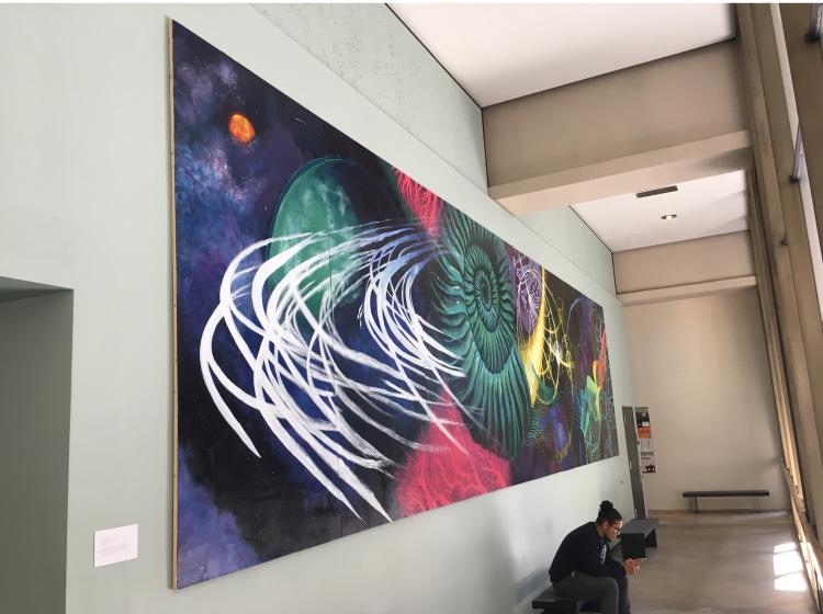 The mural is shown where it is installed in a hallway in Science Building 1, seen from the far left end. The mural features scientific imagery including a seashell-like shape surrounded by rings like Saturn. There is a work table in the foreground, with paints and brushes. There are benches in front of the wall below the mural, and a student sits on one.