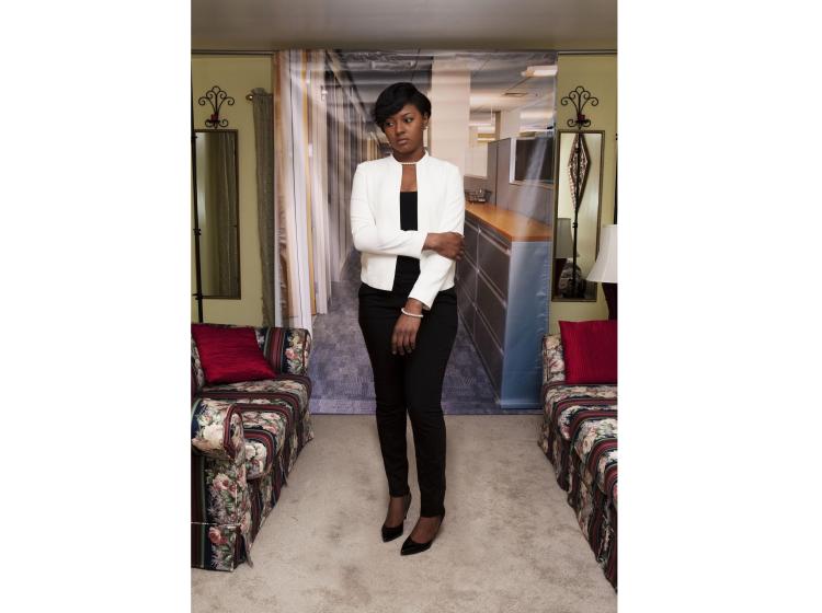 A young Black woman stands in front of a photo backdrop of a generic office suite. Her pose appears apprehensive. The backdrop is hung in a home setting, with couches, a lamp and wall mirrors visible.