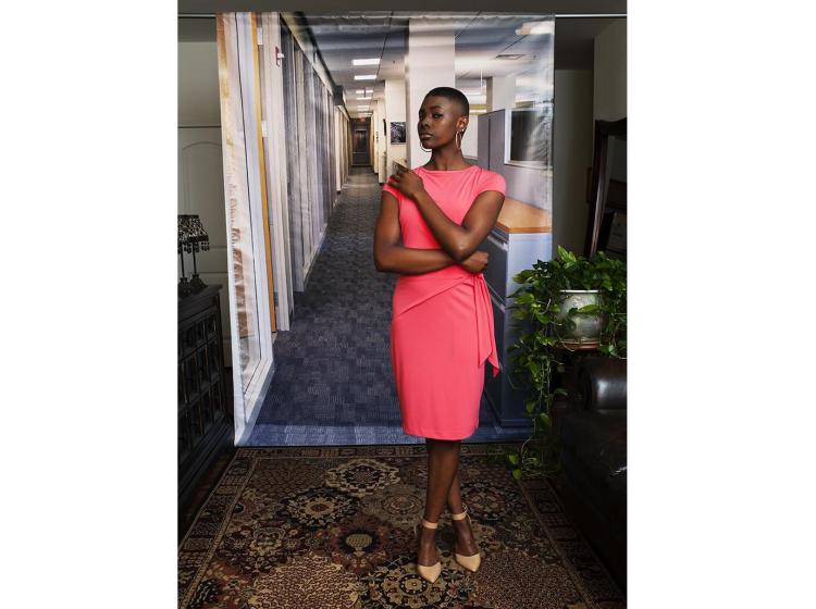 A young Black woman stands in front of a photo backdrop of a generic office suite. Her pose is confident, arms crossed in front of her. In front of and around the backdrop are carpet, furniture and a potted plant.