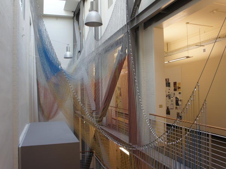 Suspended in a light well is a flowing, delicate structure of strings of colored beads that hang from two cables, in a hammock-like shape. The beads are yellow, red and blue. 