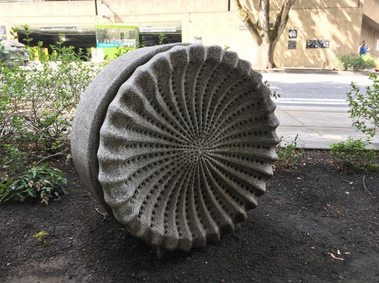 A granite sculpture sits in the dirt swale next to the sidewalk on 6th Avenue. The sculpture is shaped like a microscopic organism, somewhat resembling a wheel.