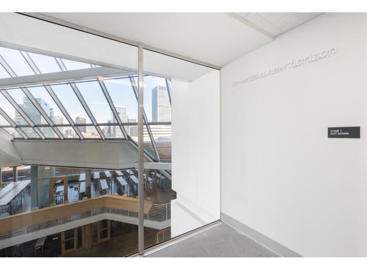 The corner of a room. At left is a floor-to-ceiling window. High on the wall at right is a line of text, in white raised italic lettering. It reads, "to write a new horizon."